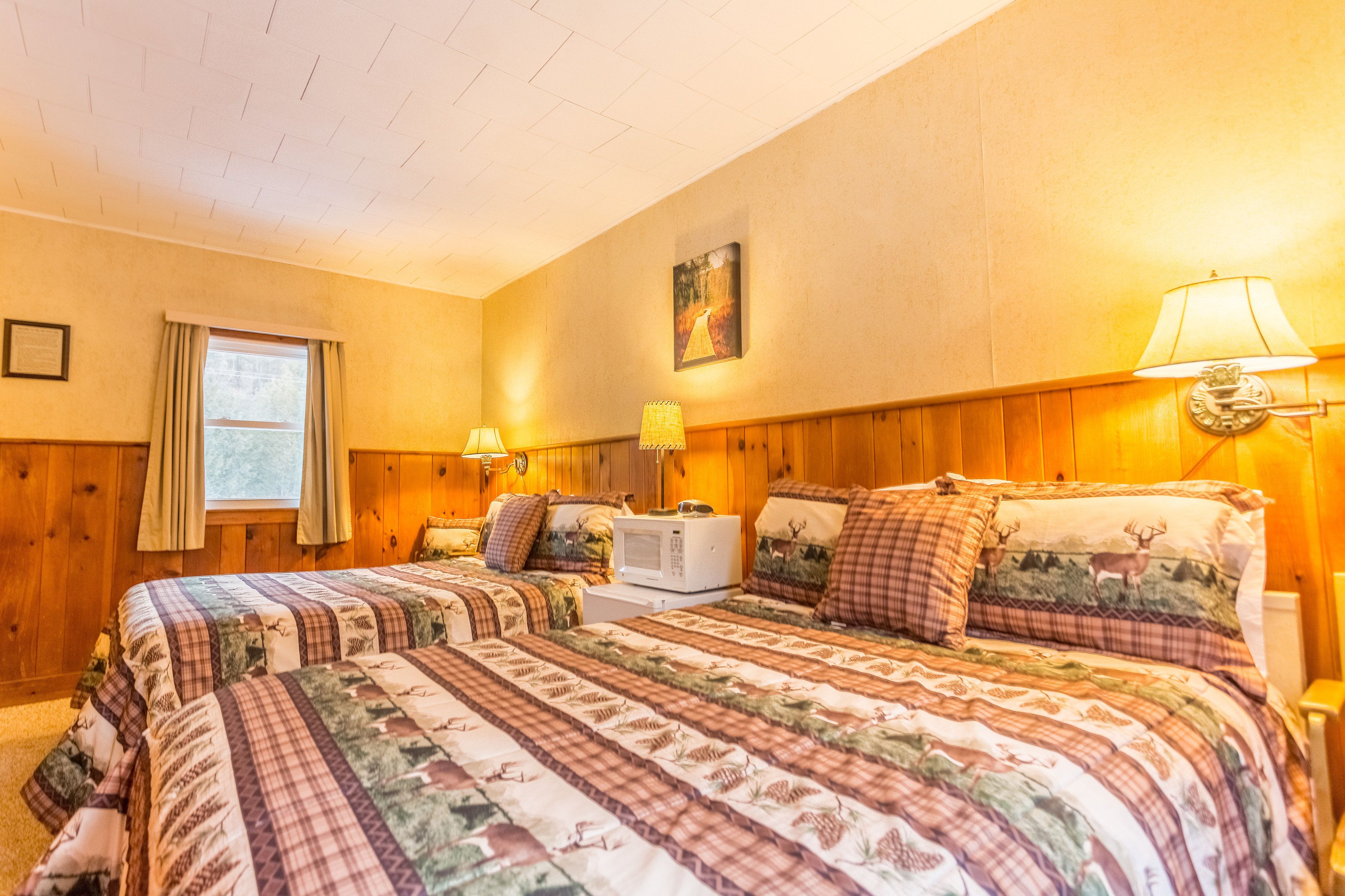 A cozy bedroom with two beds, wooden paneling, a window with curtains, and wall-mounted lamps.