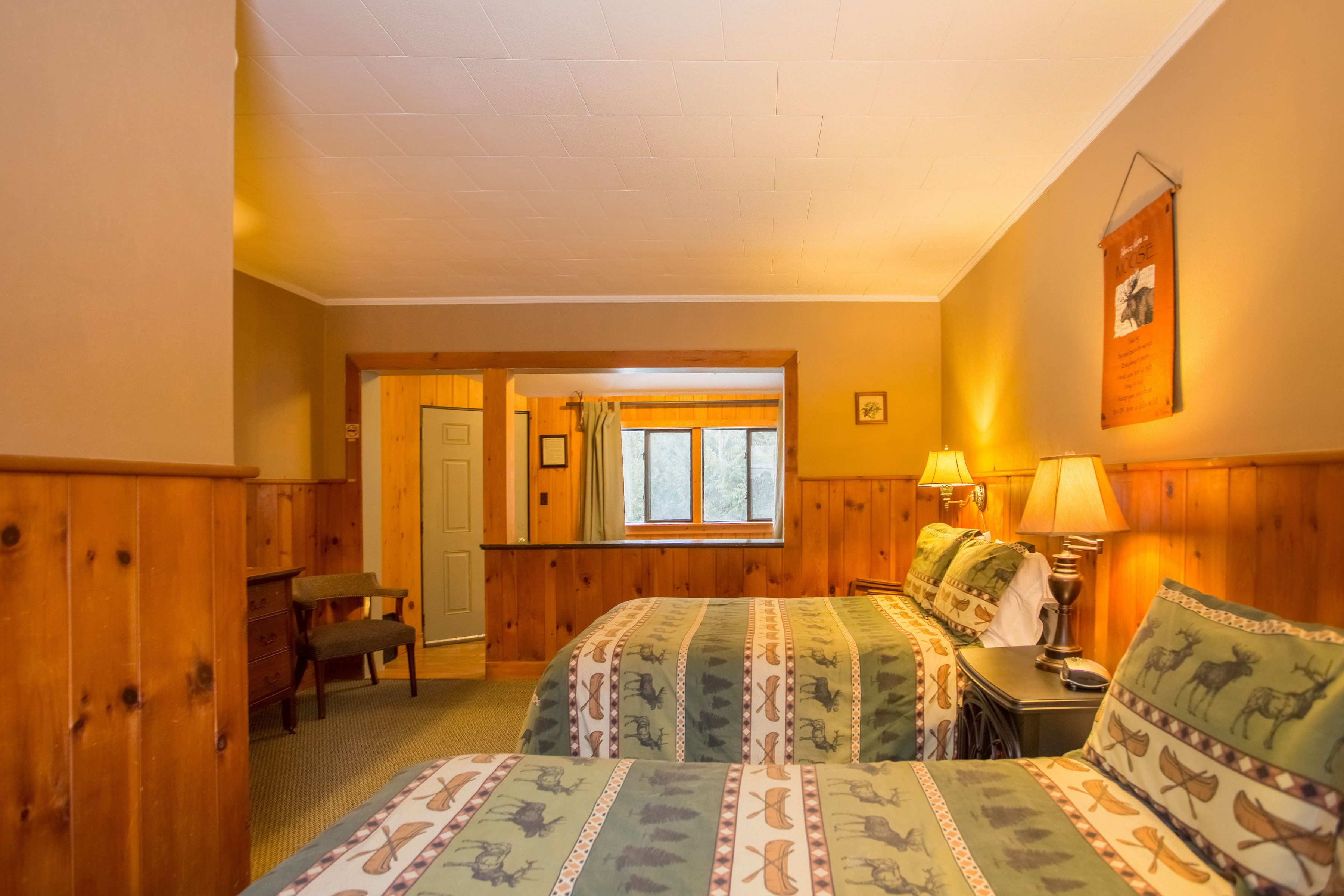 A cozy room with wooden paneling, a bed with patterned bedding, a desk with a lamp, and a window with curtains.