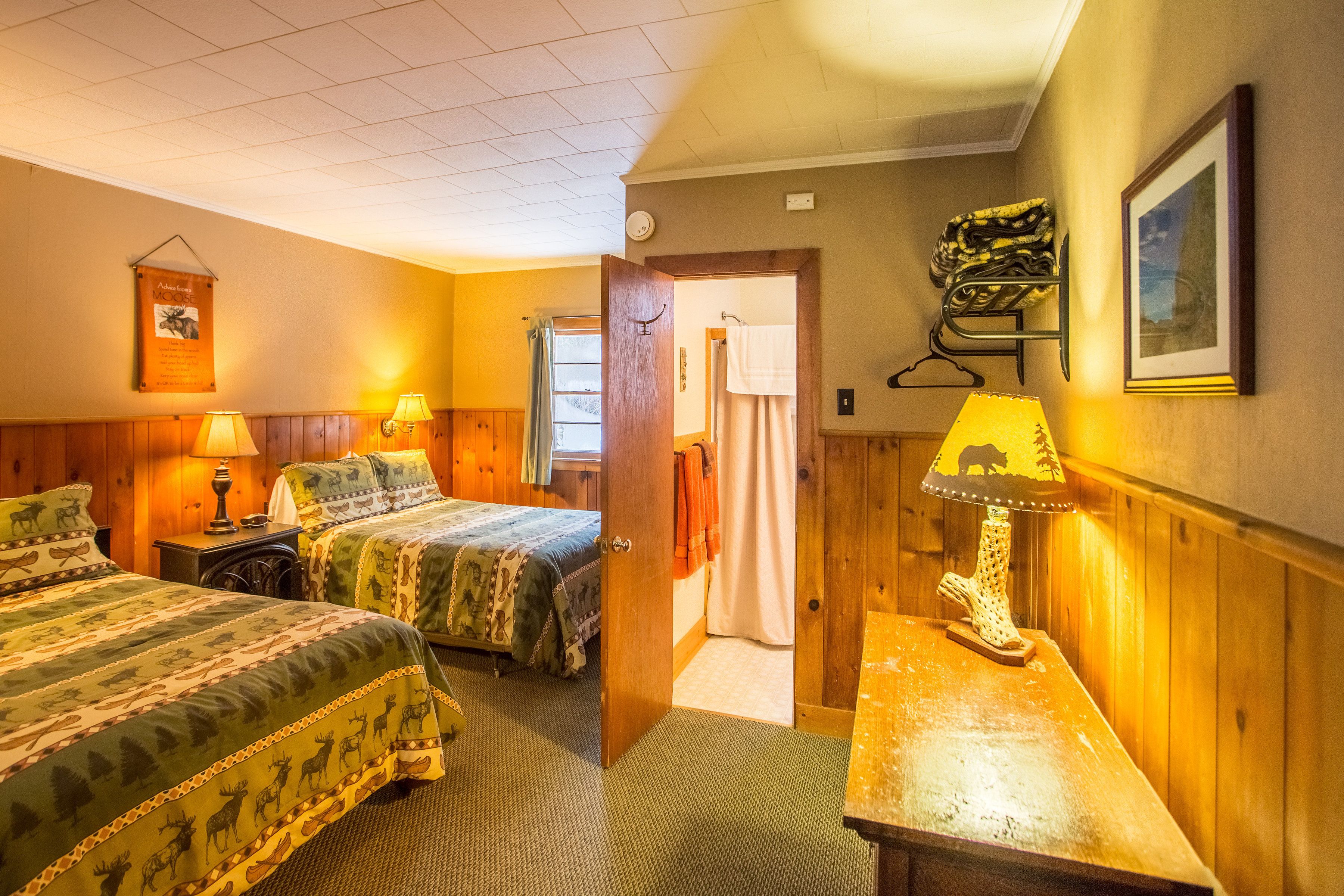 A hotel room with two beds, wooden walls, a wooden desk with a lamp, a framed picture, and a door leading to another room.