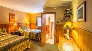 A hotel room with two beds, wooden walls, a wooden desk with a lamp, a framed picture, and a door leading to another room.