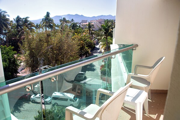 A balcony with a glass railing overlooking a parking area, palm trees, and mountains in the distance.