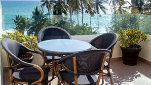 A balcony with wicker chairs and a table, overlooking the ocean and palm trees.