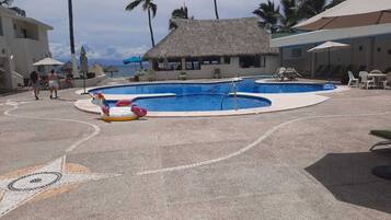 Un espace piscine comprenant deux piscines, des chaises longues, des parasols et une structure dotée d’un toit de chaume.