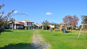 Un sendero verde que conduce a un parque infantil con tobogán y columpios, rodeado de casas con techos de tejas rojas.