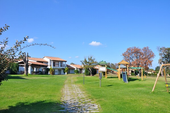 A grassy pathway leading to a playground with a slide and swings, surrounded by houses with red-tiled roofs. - Liscia Eldi Residence (San Teodoro)