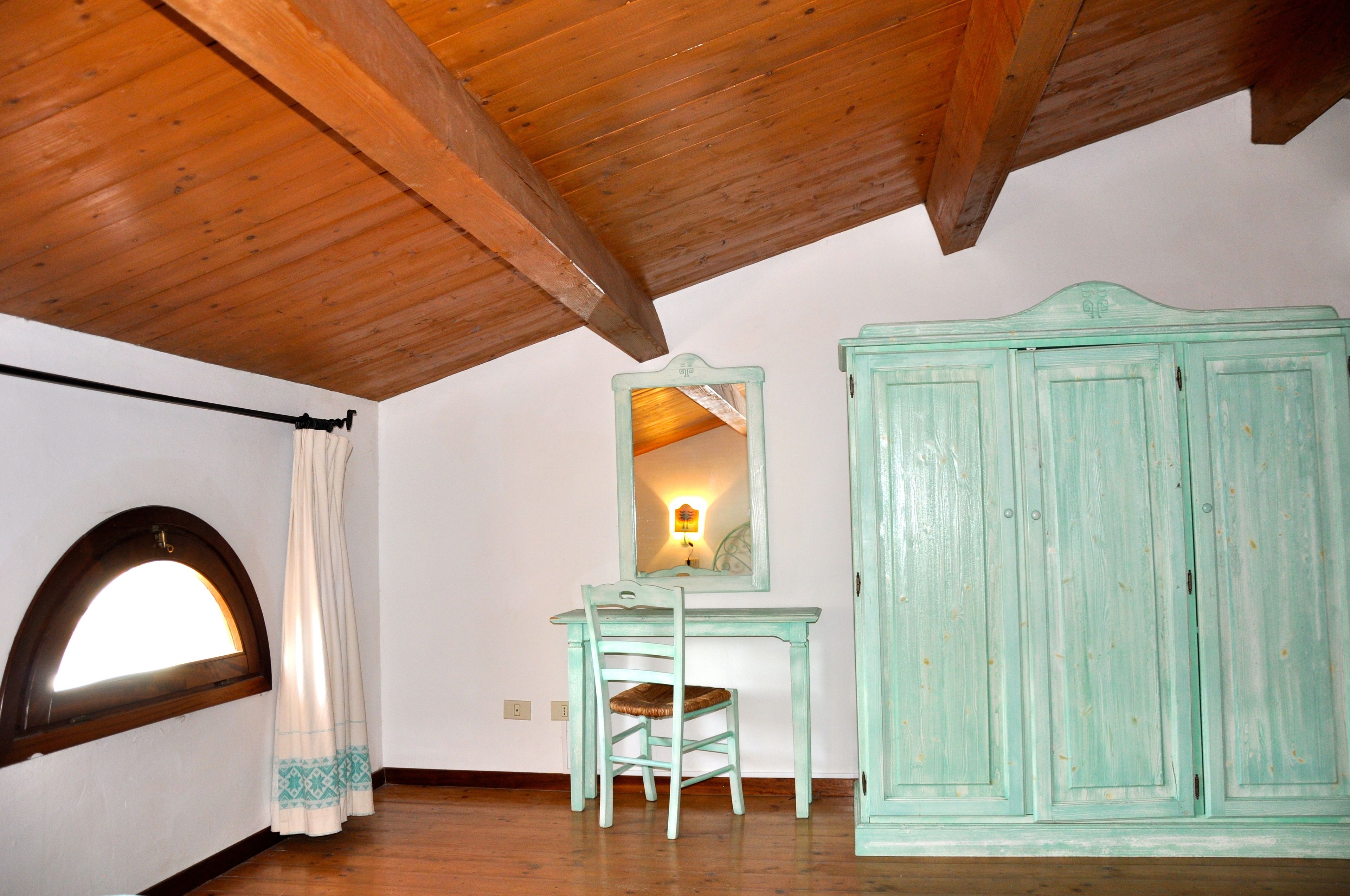 A room with a wooden ceiling, a desk with a chair, a large cabinet, and a window with a curtain.