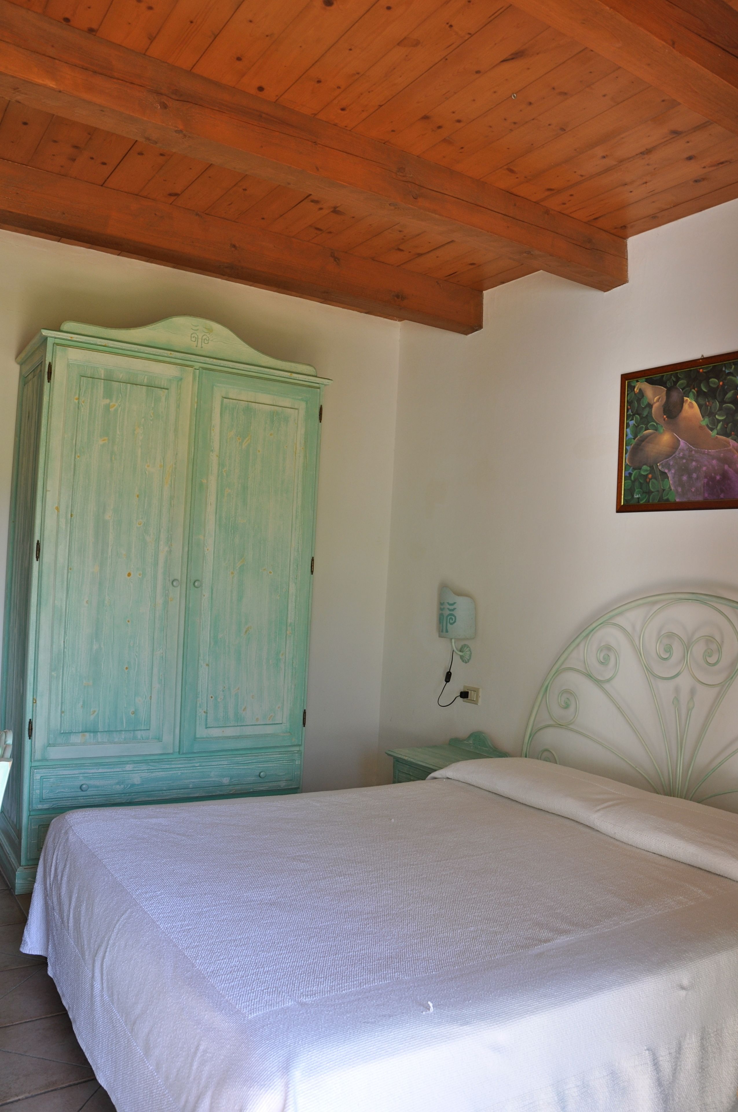 A bedroom with a wooden ceiling, a green wardrobe, a white bed, and a wall-mounted nightstand with a lamp.