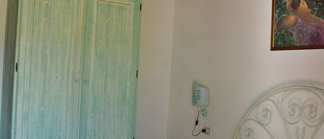 A bedroom with a wooden ceiling, a green wardrobe, a white bed, and a wall-mounted nightstand with a lamp.
