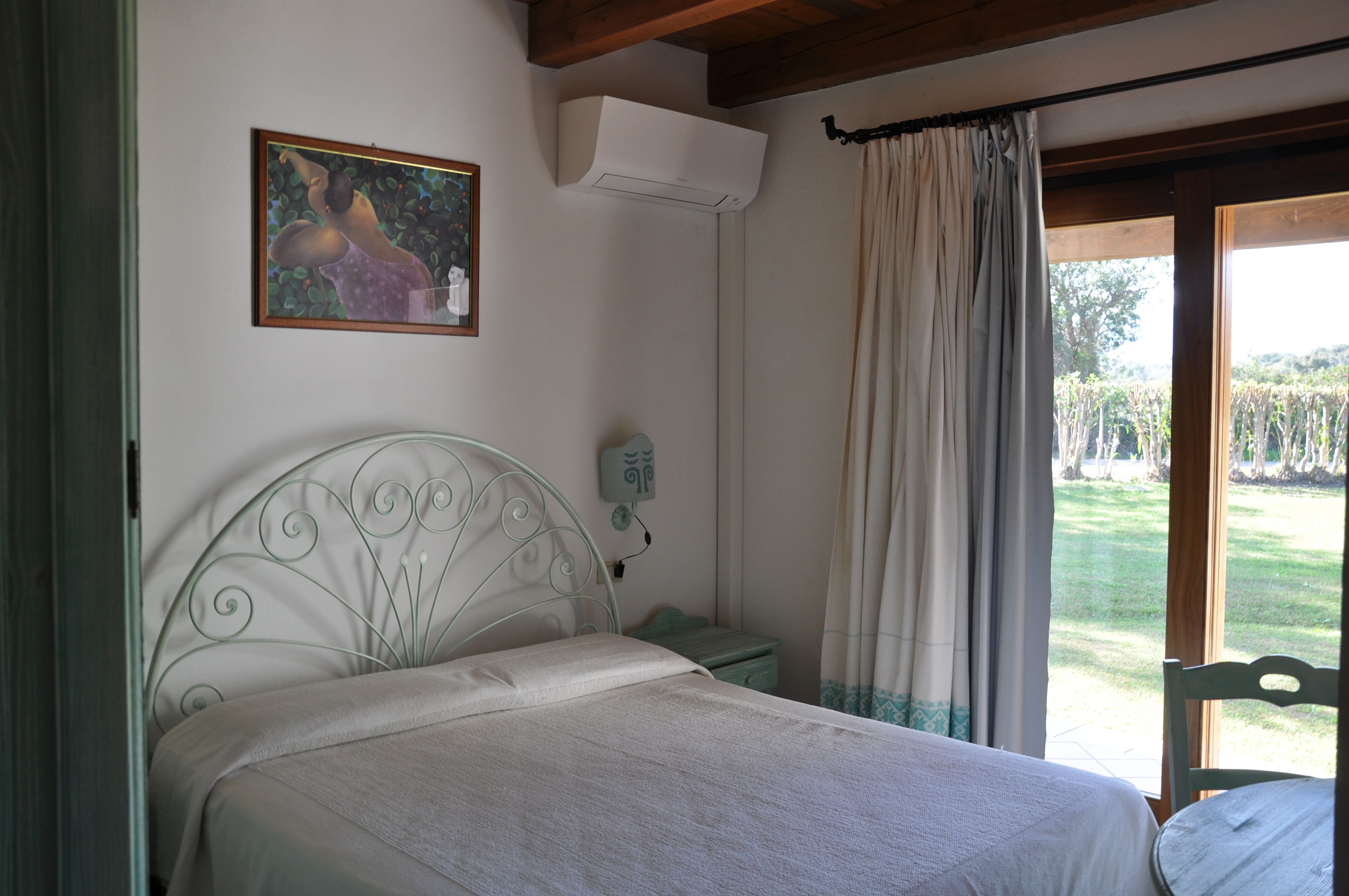 Een slaapkamer met een metalen bed, een nachtkastje, een airconditioning, een telefoon aan de muur en uitzicht op een tuin door een raam met gordijnen.