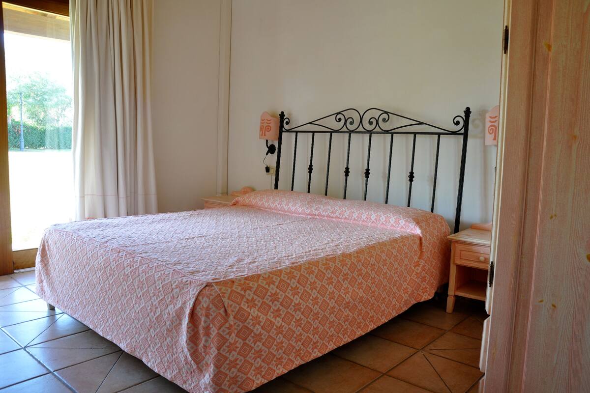 Un lit simple avec une couvre-lit à motifs, dans une pièce dont le sol est carrelé et qui possède une fenêtre avec des rideaux.
