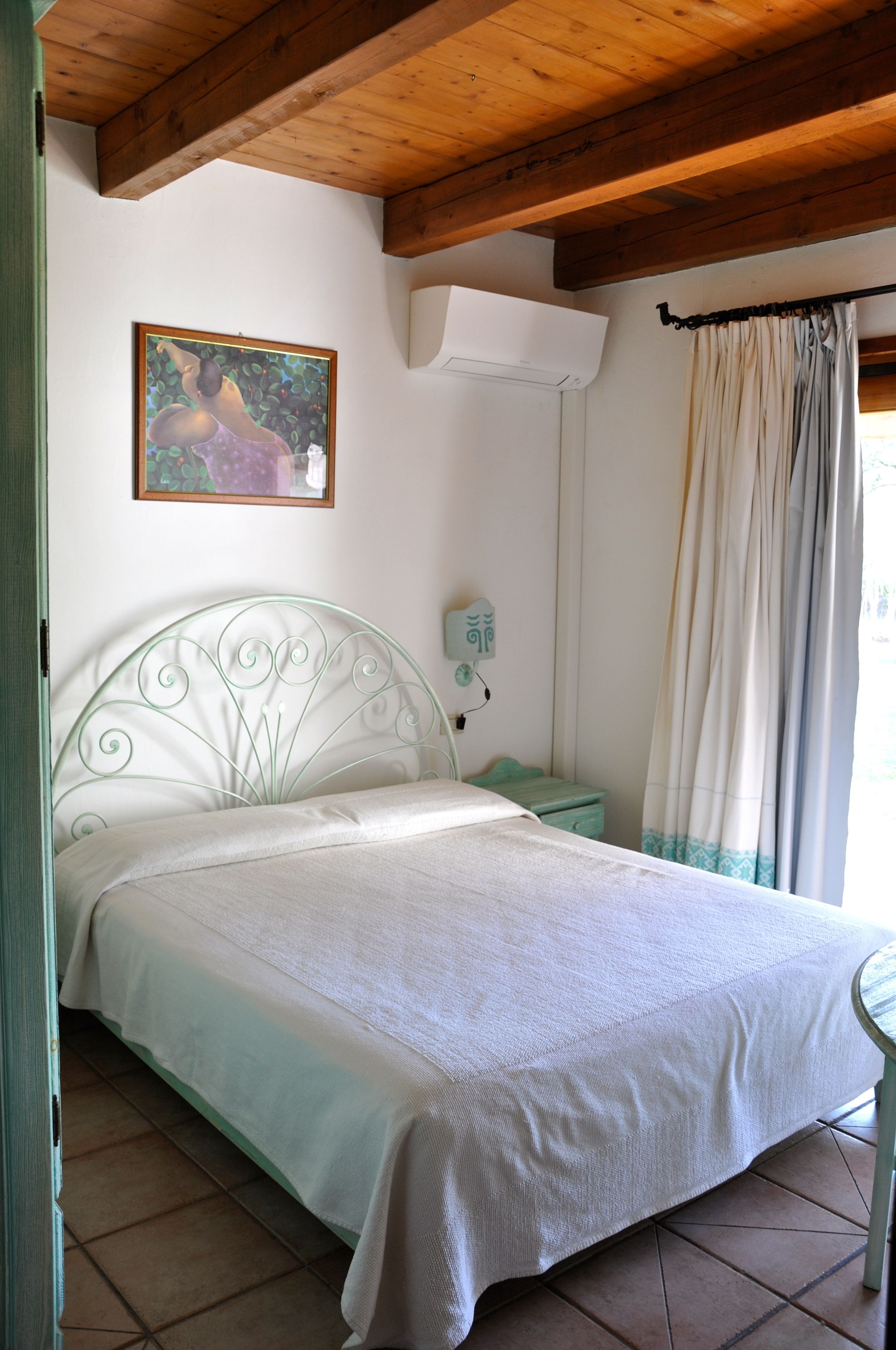 Un dormitorio con estructura metálica para la cama, una mesita de noche, aire acondicionado de pared y una ventana con cortinas.