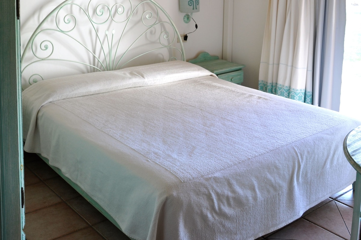 A bedroom with a metal bed frame, a bedside table, a wall-mounted air conditioner, and a window with curtains.