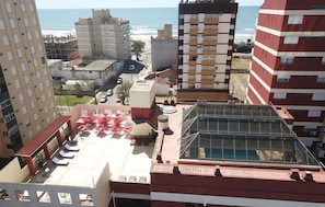 Terraço no telhado com estrutura de vidro, cercado por edifícios altos, com vista para uma praia.