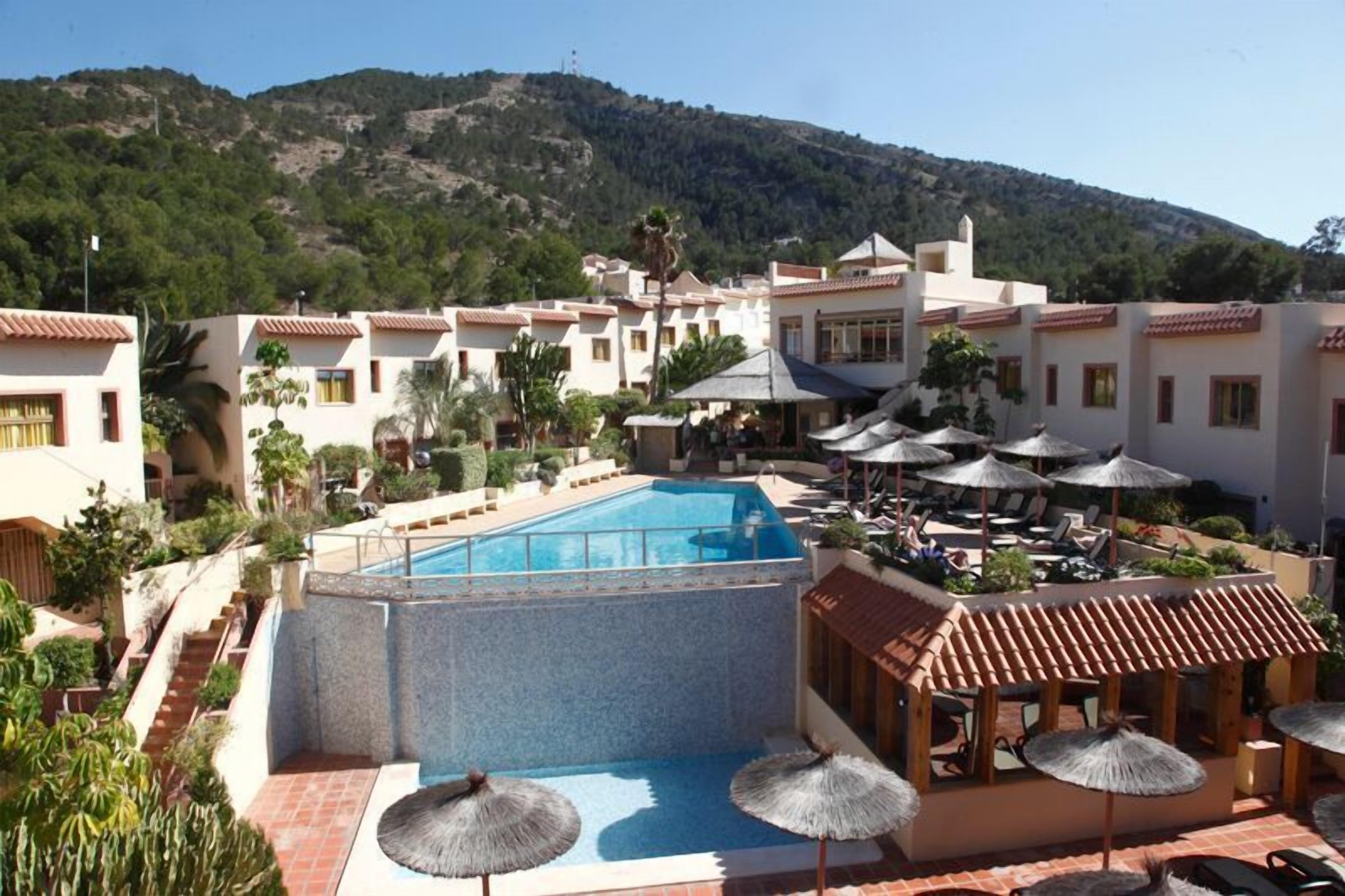 A hotel with a swimming pool, outdoor seating with umbrellas, and a mountainous backdrop.