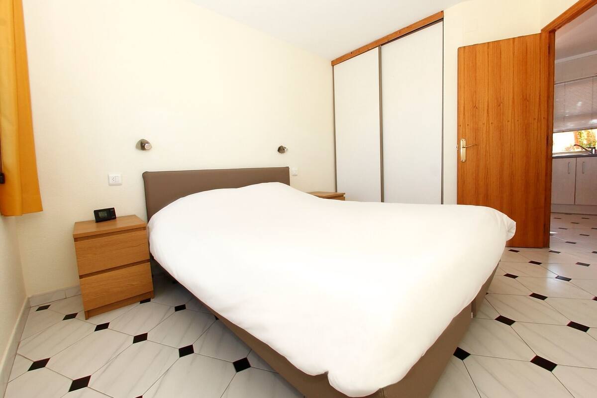 A bedroom with a bed, a nightstand, a wooden wardrobe, and a door leading to a bathroom.