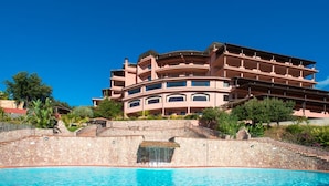 Un hotel de varios pisos con una piscina al frente, rodeado de vegetación y un cielo azul despejado.