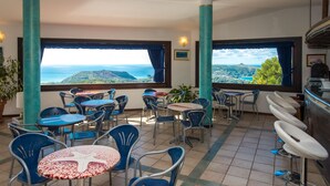 Una terraza con mesas y sillas, ventanas amplias con cortinas y vista al mar.