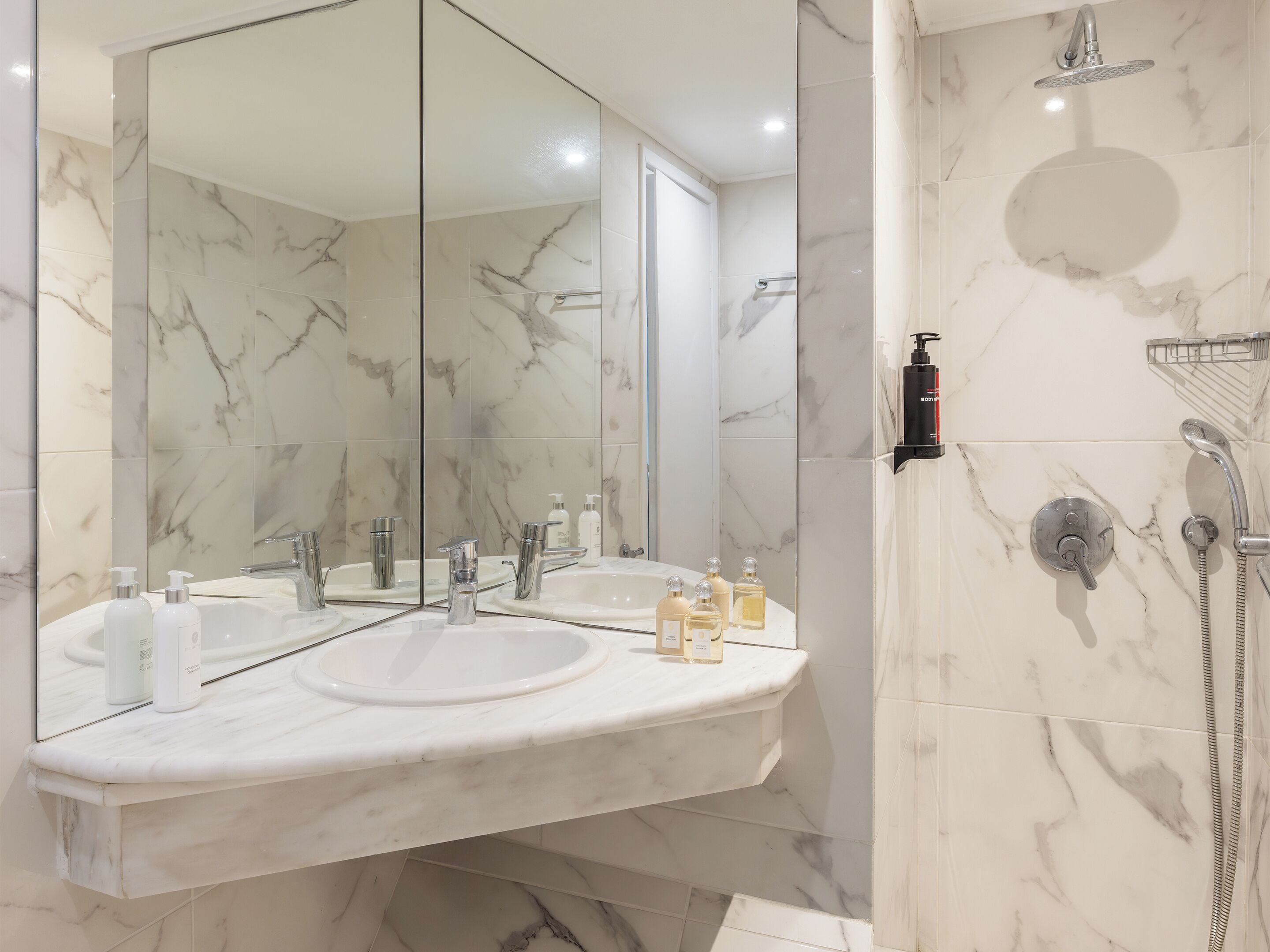 A bathroom with a marble vanity, a large mirror, a shower with a handheld showerhead, and a bottle of lotion on the sink.