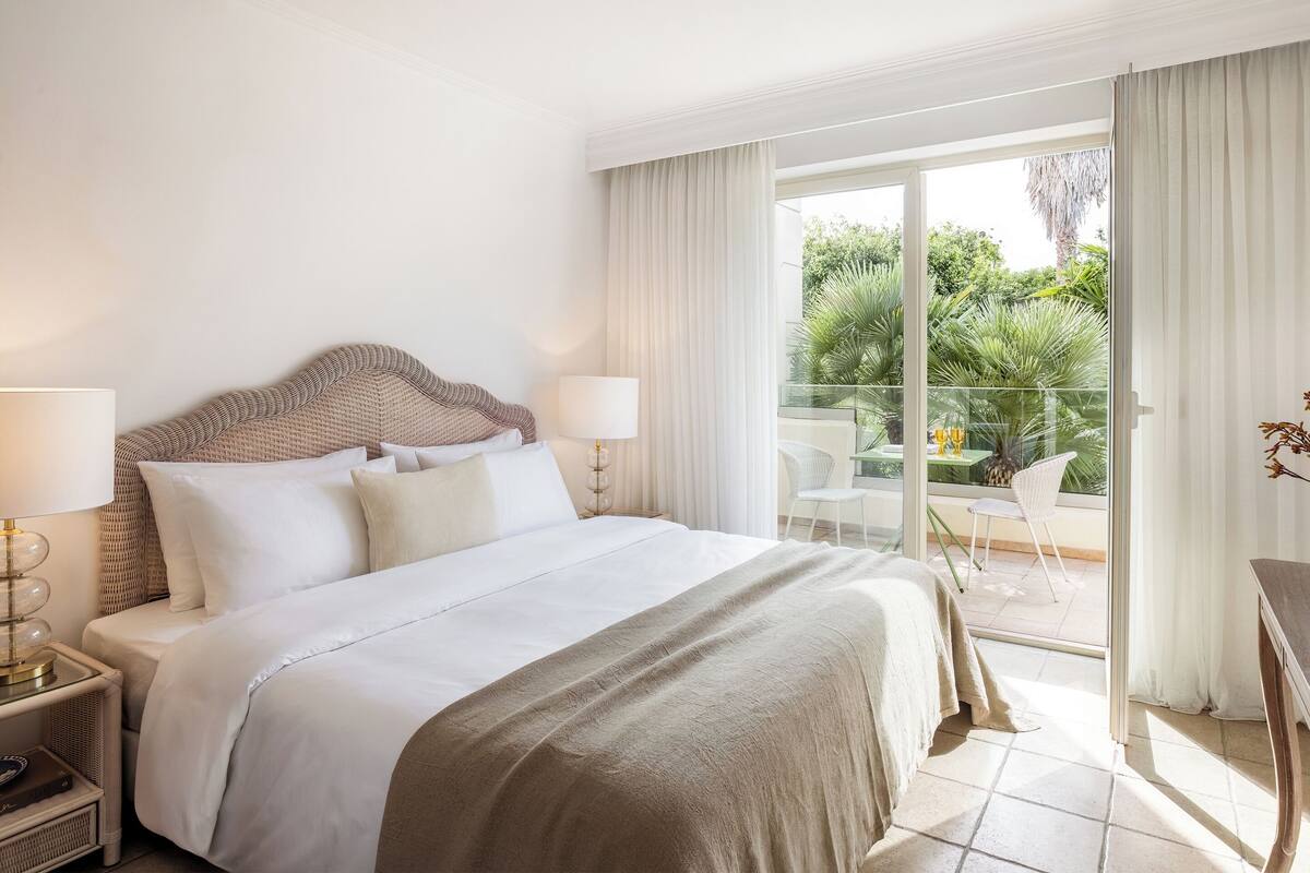 Une chambre d’hôtel avec un grand lit, deux tables de chevet avec des lampes, un balcon avec une table et des chaises, et une vue sur la verdure environnante.