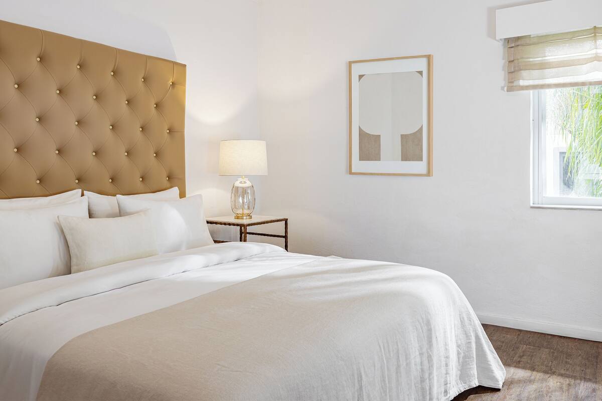 Une chambre d’hôtel avec une tête de lit capitonnée, une table de chevet avec une lampe, une œuvre d’art encadrée au mur et une fenêtre avec des stores.