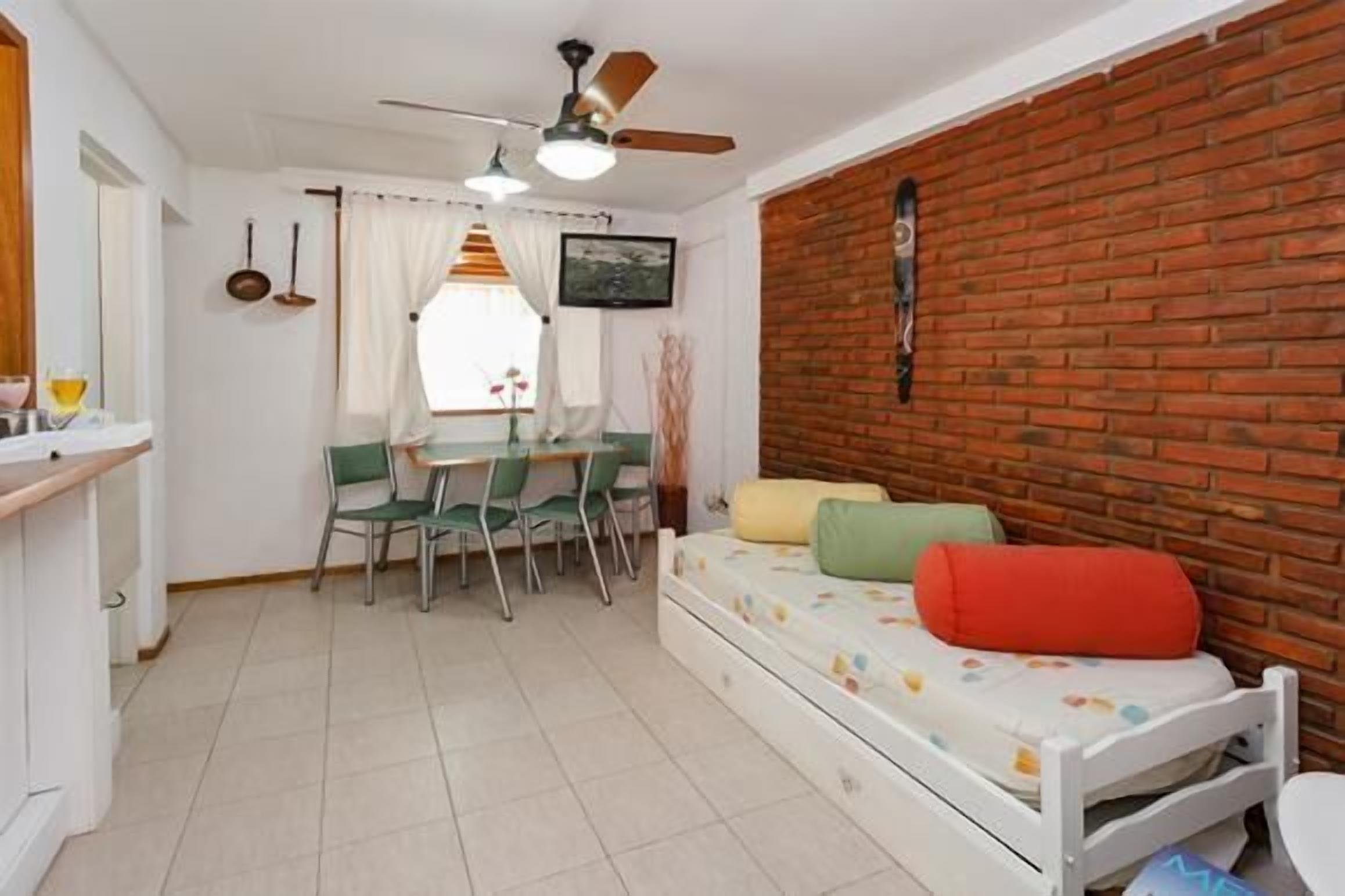 A compact room with a bed, a dining table with chairs, a ceiling fan, and a brick wall.