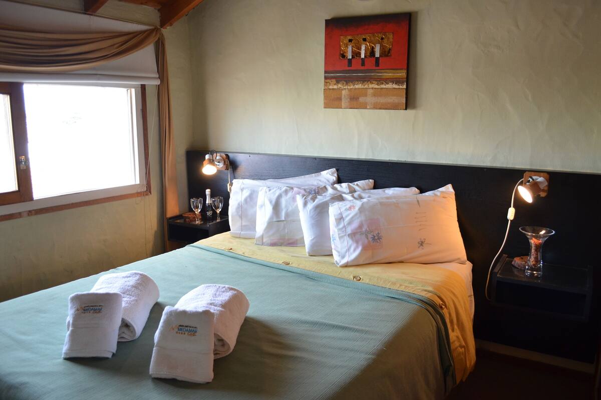 A neatly made bed with white linens and towels, a bedside table with a lamp, and a painting on the wall.