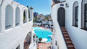 Une vue sur une piscine entourée de bâtiments blancs dotés de fenêtres en arc de cercle et d’un escalier menant à une porte.