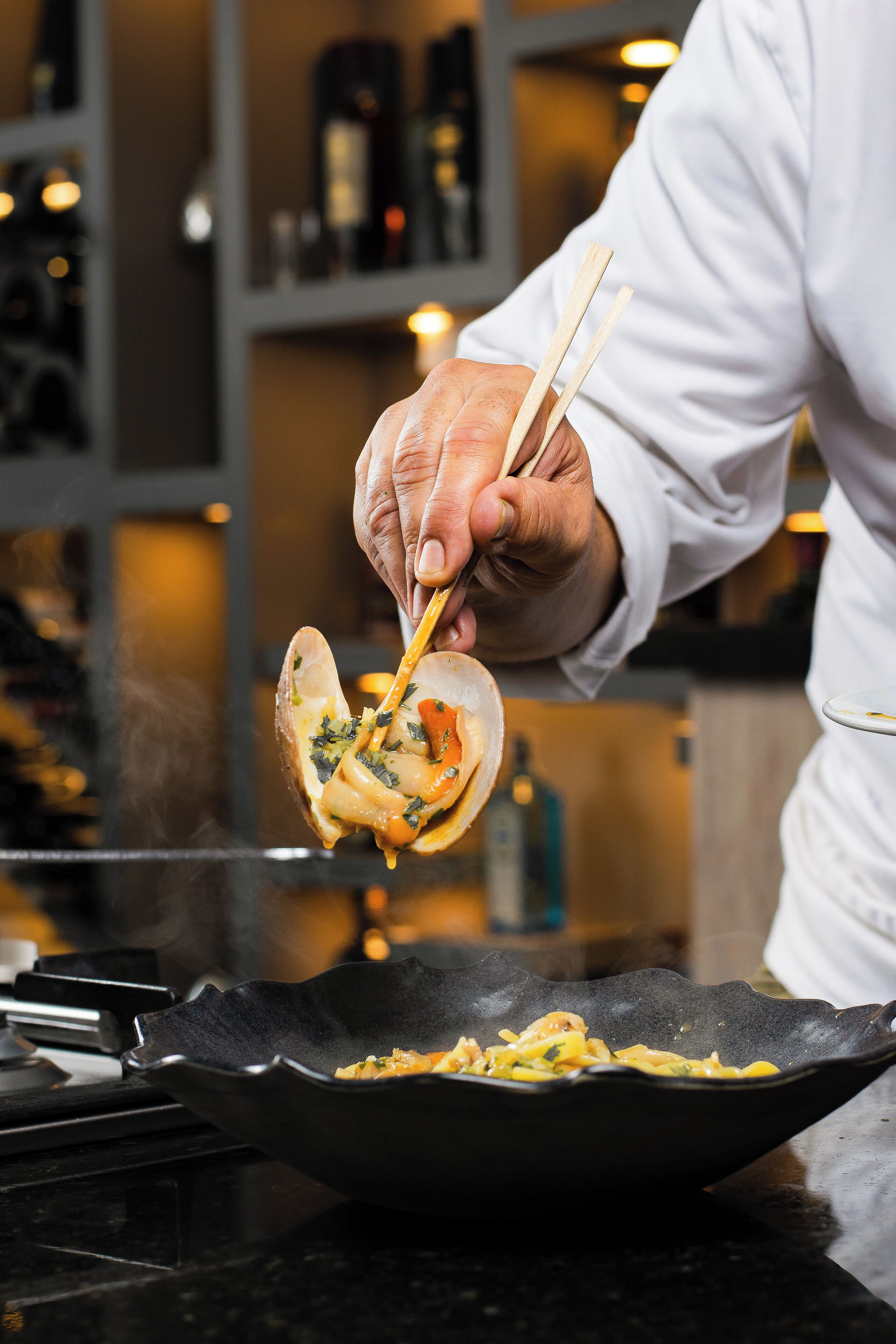 A chef holding a bowl with food, a pan on the counter, and shelves with bottles in the background.