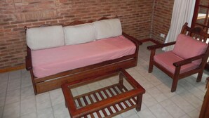 Una habitación con paredes de ladrillo, un sofá y una silla de madera, una pequeña mesa y dos cuadros en la pared.