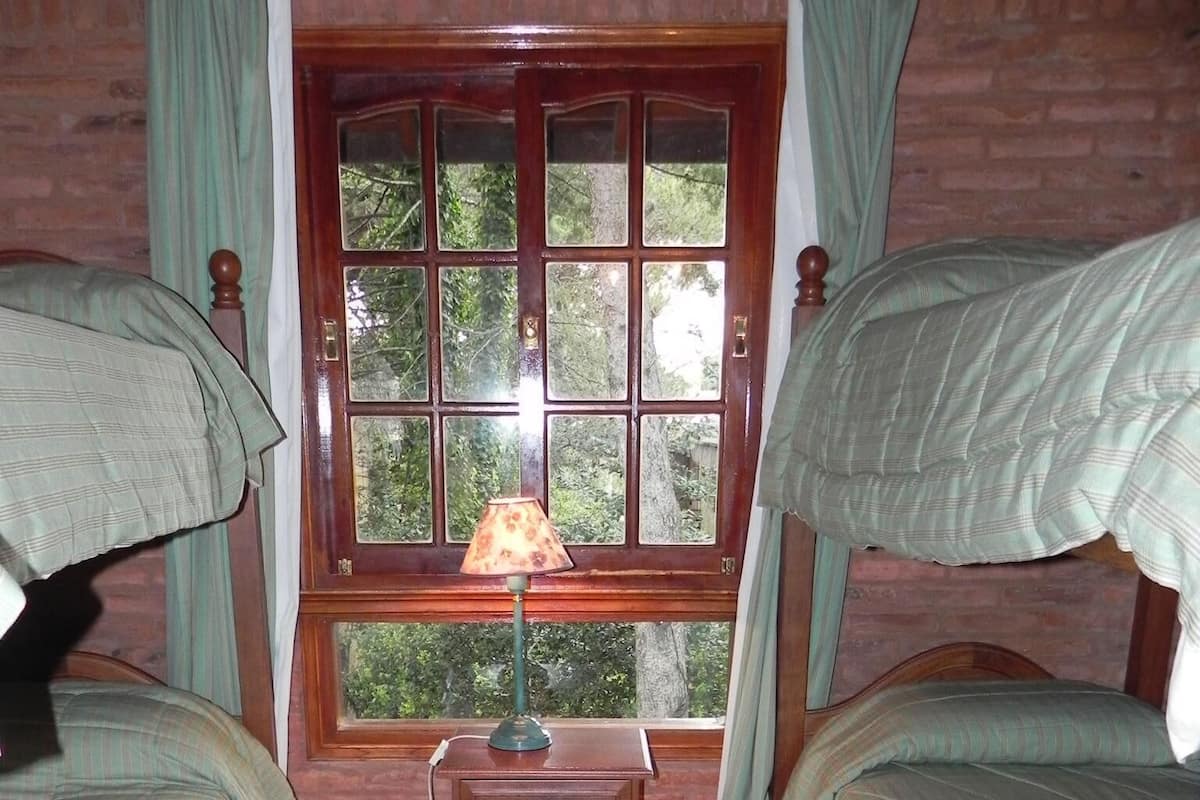 Una habitación con dos camas, una mesita de noche, una ventana con cortinas y una lámpara.