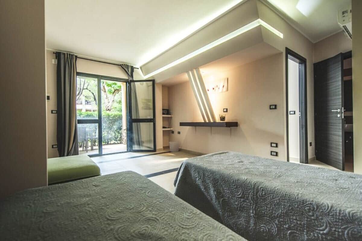 A hotel room with two beds, a sliding glass door to the outside, a small table, and a shelf with decorative items.