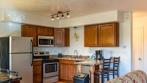 A compact kitchen with wooden cabinets, a stainless steel refrigerator, a microwave, an oven, and a coffee maker. There is a trash can and a patterned sofa in the room. - Crown Resorts at Silverwoods (DuBois)