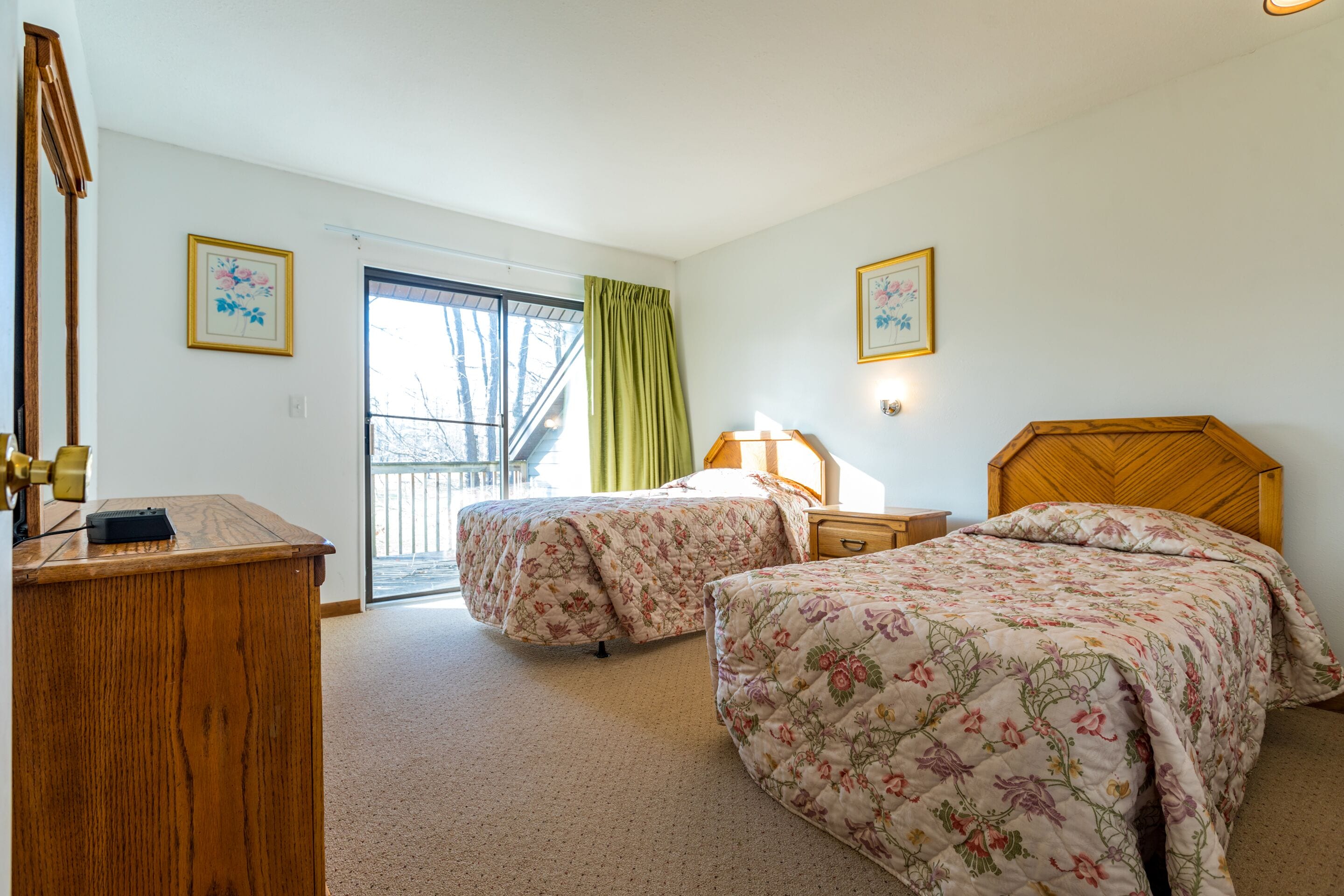 A bedroom with two beds, a wooden desk, a lamp, and a window with a view of trees.