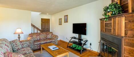 A living room with a sofa set, a coffee table, a flat-screen TV, a fireplace, and a staircase in the background.
