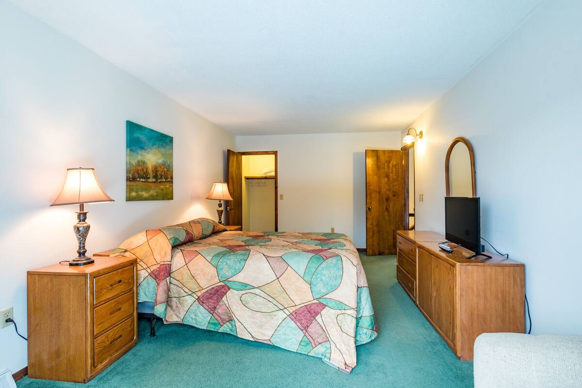 Een slaapkamer met een bed, twee nachtkastjes met lampen, een kapstok en een televisie, en een deur naar een andere kamer.