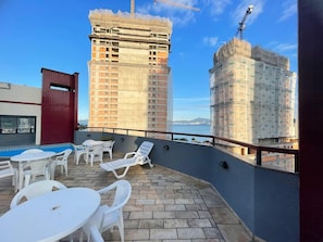 Terraço no telhado com mobiliário de plástico branco, piscina e vista para um horizonte urbano com edifícios em construção.
