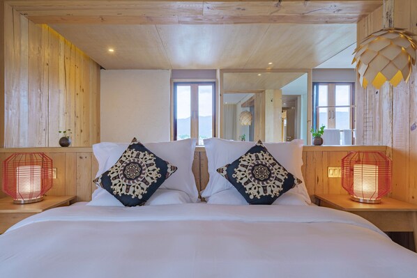 A bedroom with a wooden ceiling, two bedside tables with red lamps, a bed with white bedding and patterned pillows, and a large window with a view.