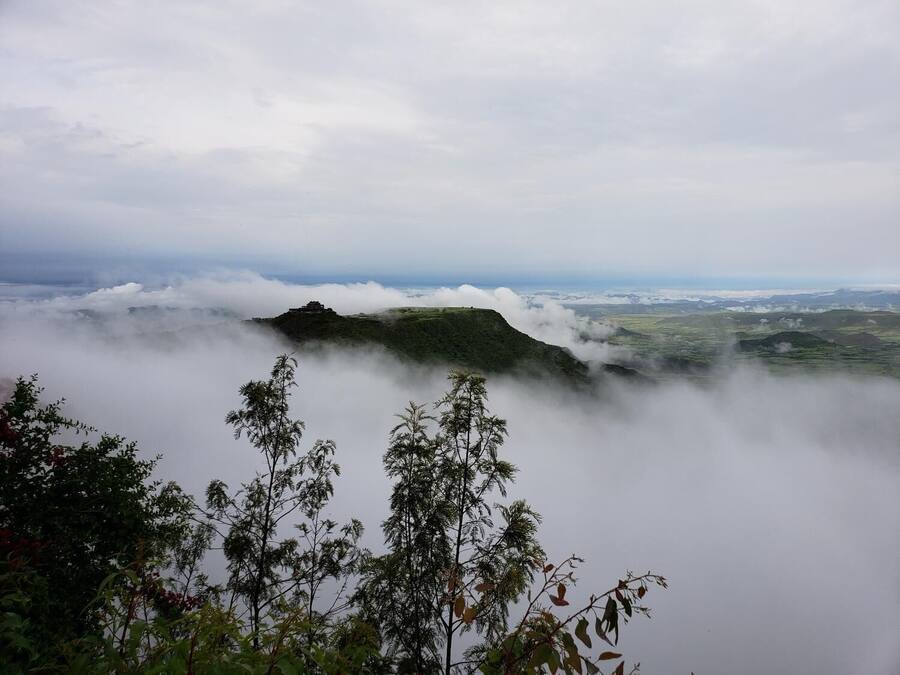 Un paisaje montañoso con una estructura en una colina, rodeada de nubes.