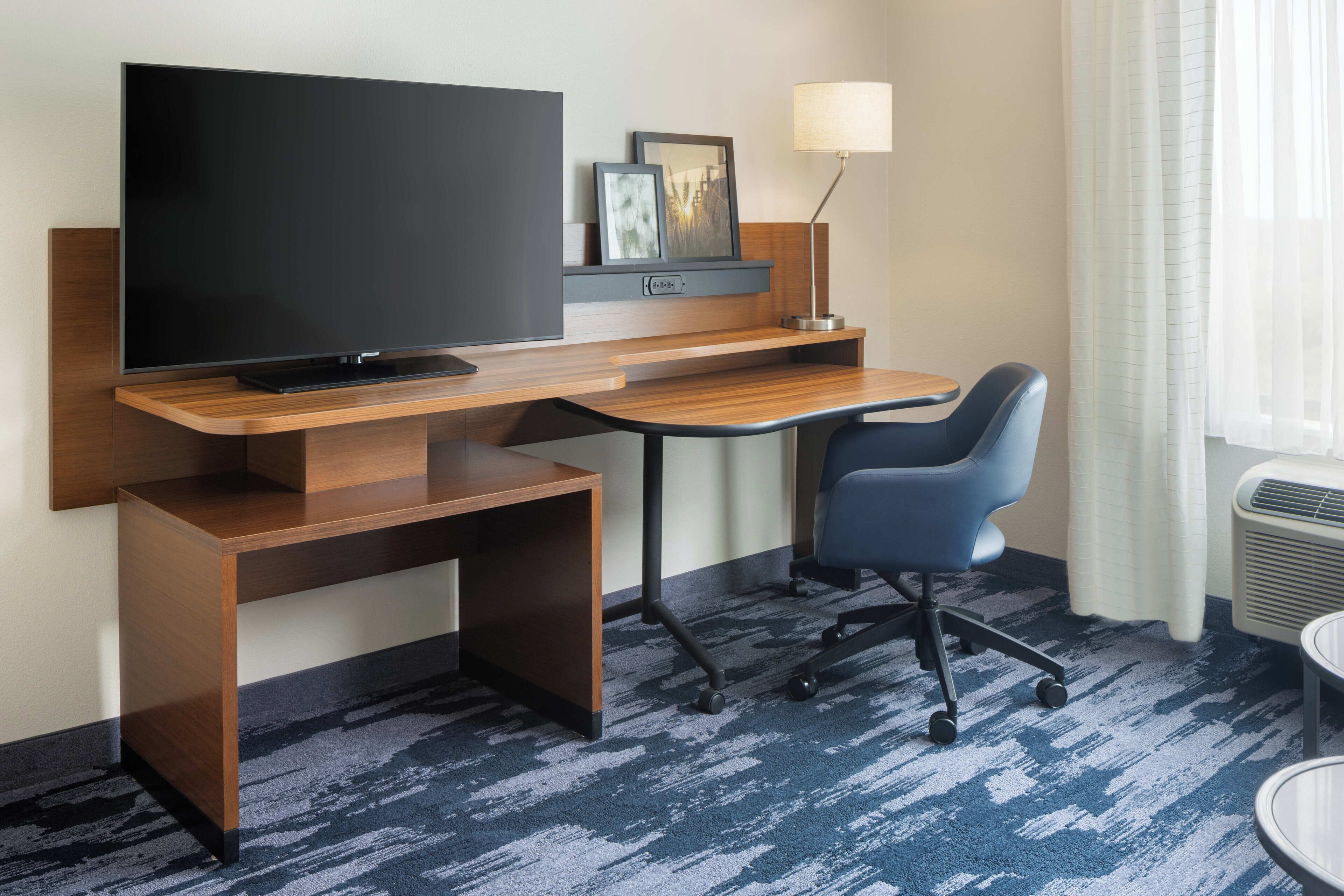 A hotel room with a wooden desk, a television, a chair, a lamp, and a window with curtains.