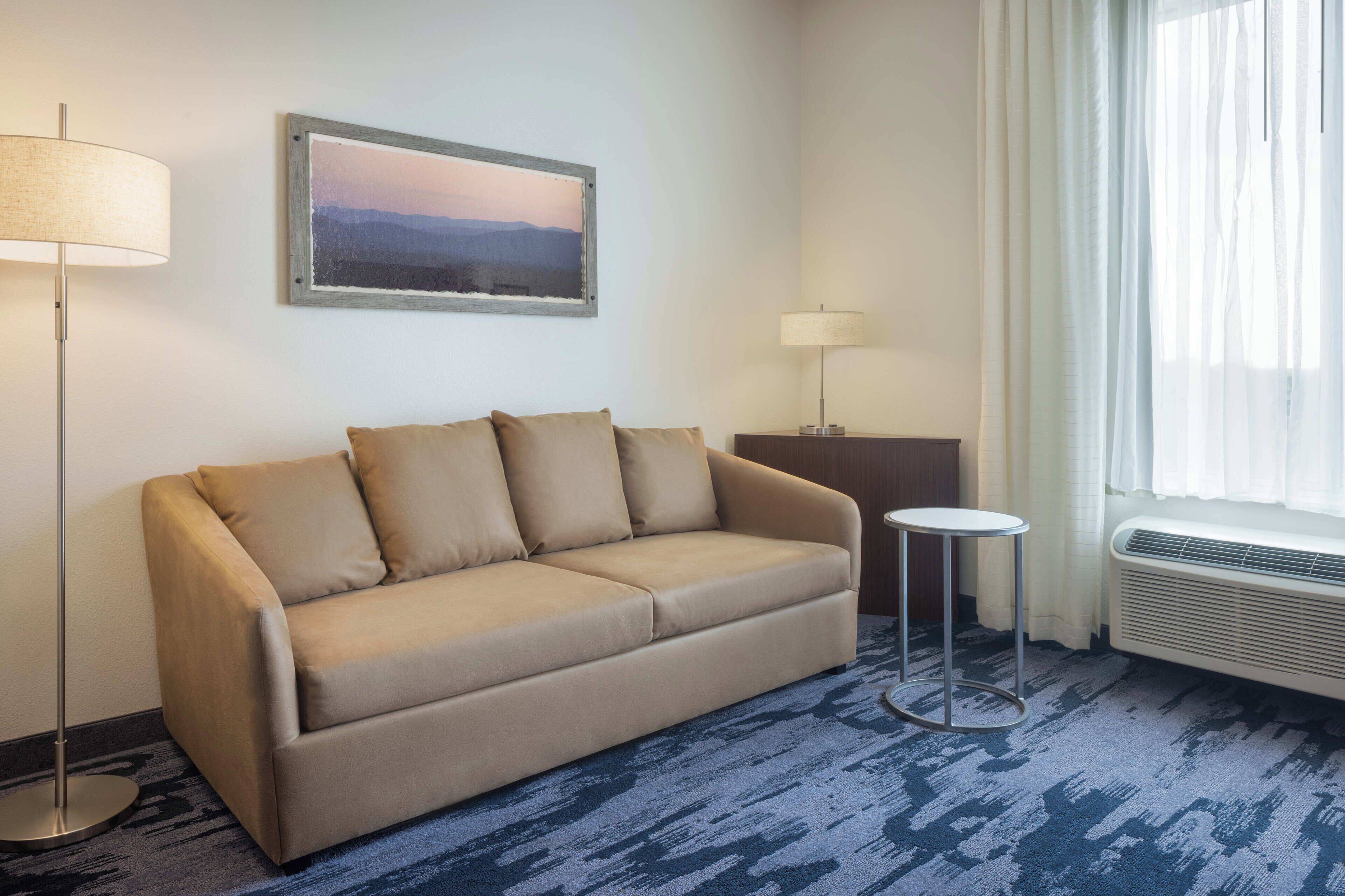 A hotel room with a beige sofa, two floor lamps, a small round table, and a large window with sheer curtains.