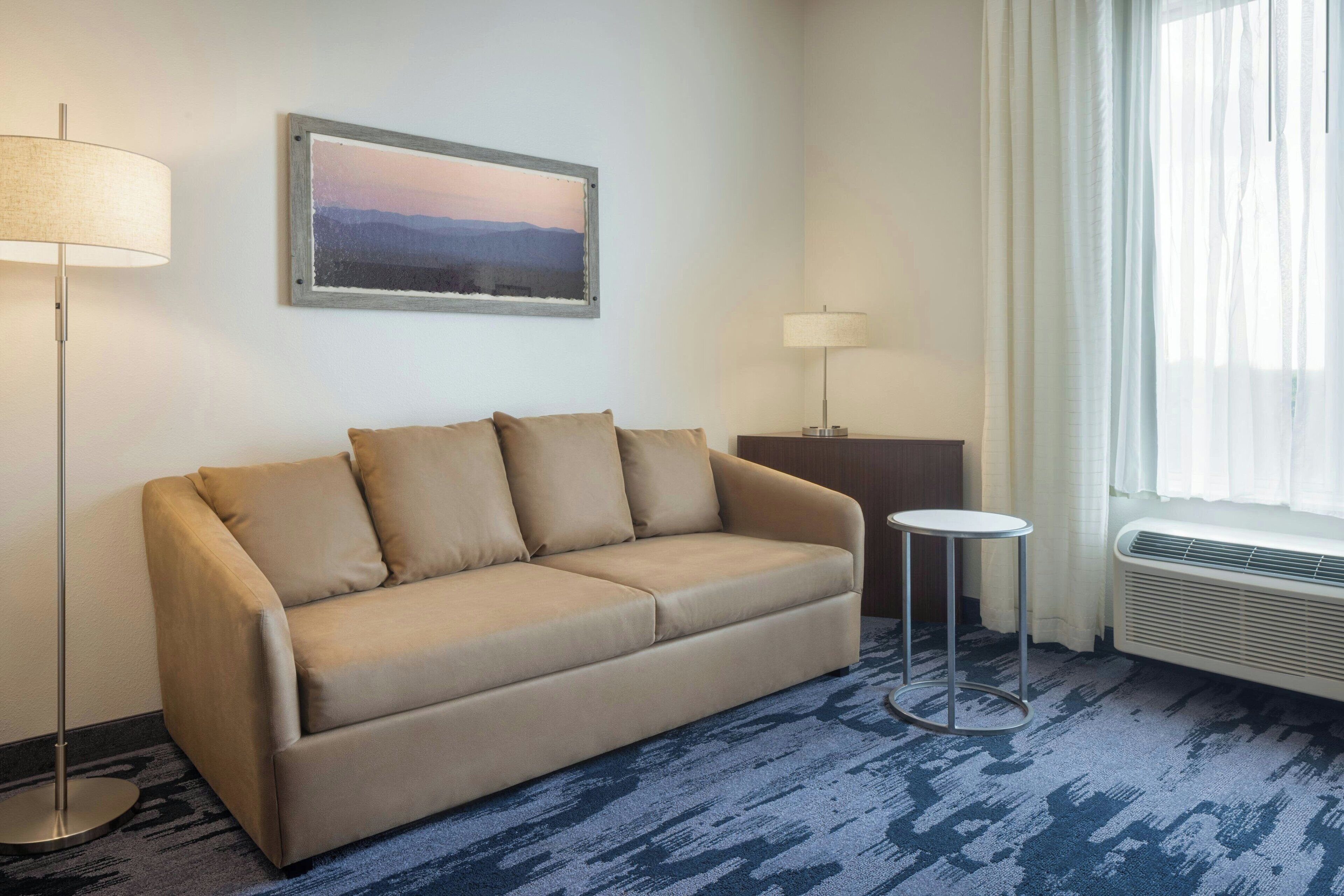 A beige sofa with four cushions, a small round table, a floor lamp, a picture on the wall, and a window with curtains.