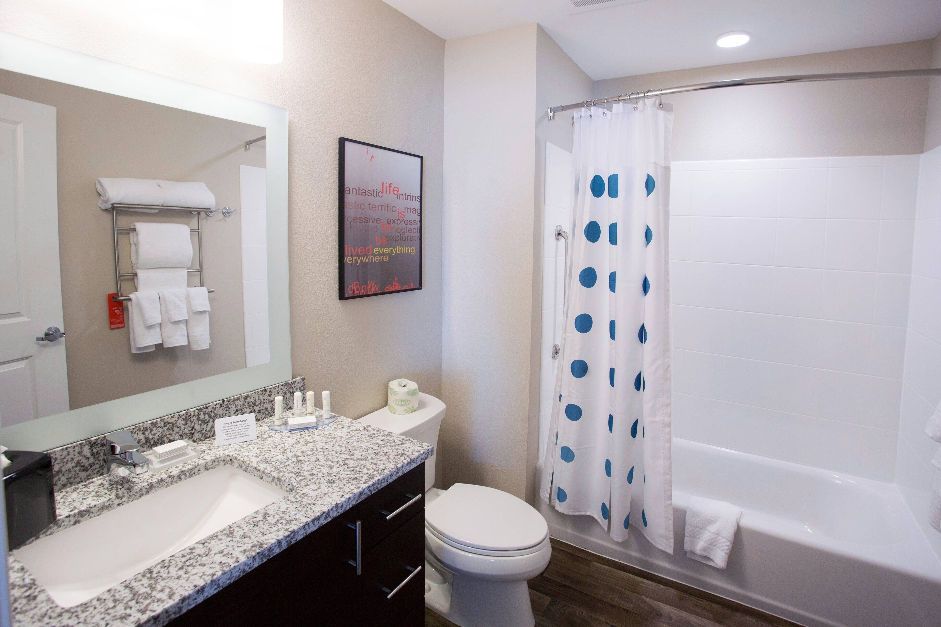 A bathroom with a sink, toilet, and bathtub. There is a towel rack with towels, a framed quote on the wall, and a shower curtain with blue polka dots.