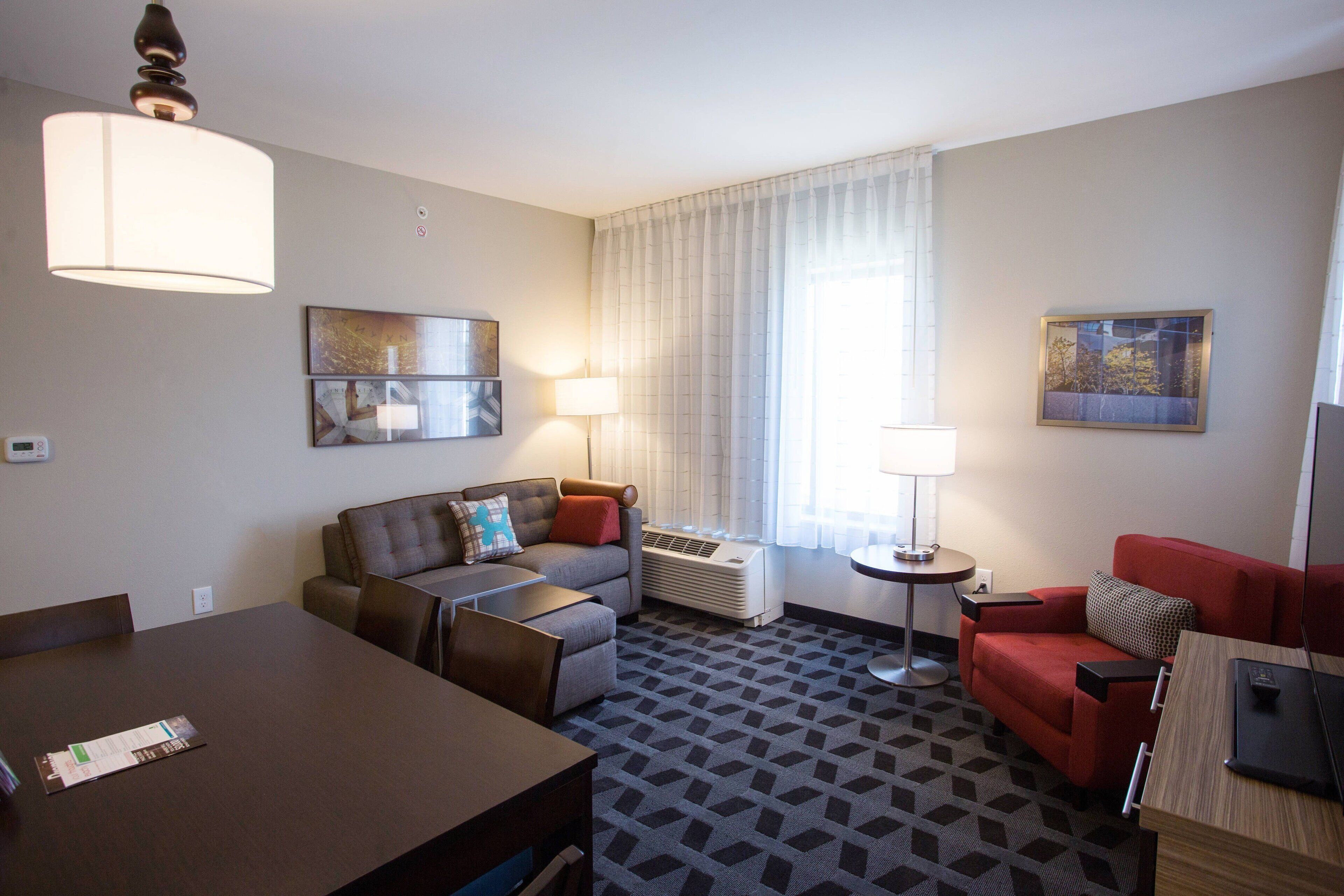 A hotel room with a dining table, two sofas, a TV, and a large window with curtains.
