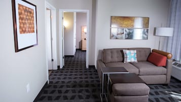 A modern hotel room with a geometric patterned carpet, a sofa with a red pillow, a small table, and a framed artwork on the wall.