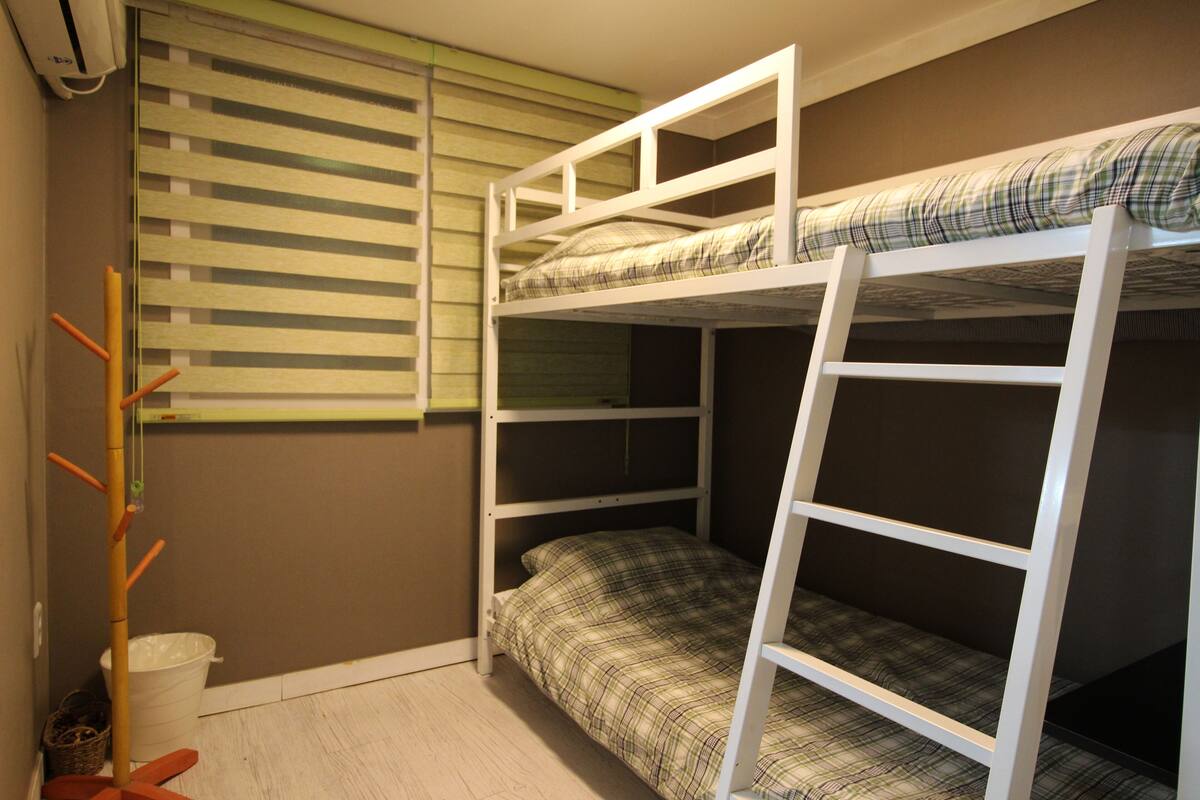 Une chambre avec un lit superposé, une fenêtre, un seau blanc et un balai.