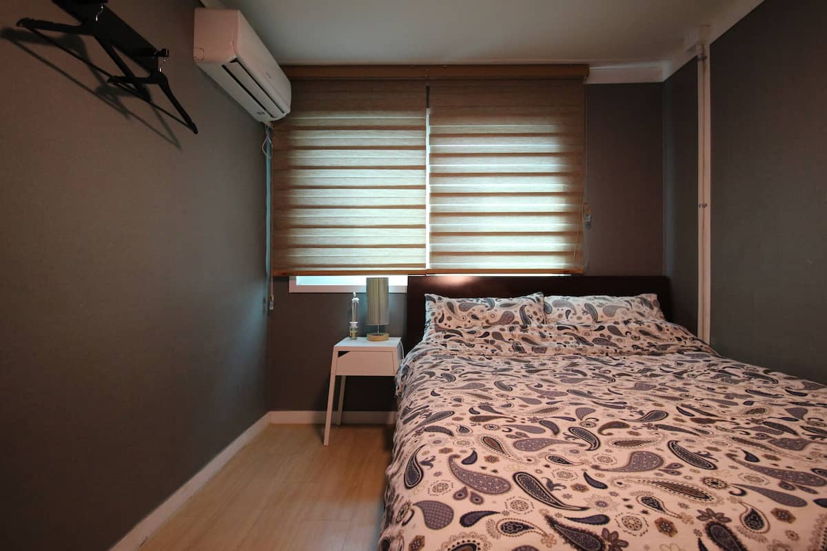 Une chambre à coucher comprenant un lit, une table de chevet, un climatiseur et une fenêtre avec des stores.