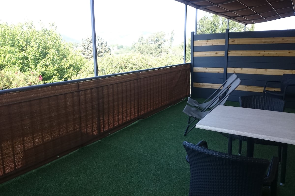 Um pátio externo com mesa e cadeiras, cerca de madeira e vista para o verde.