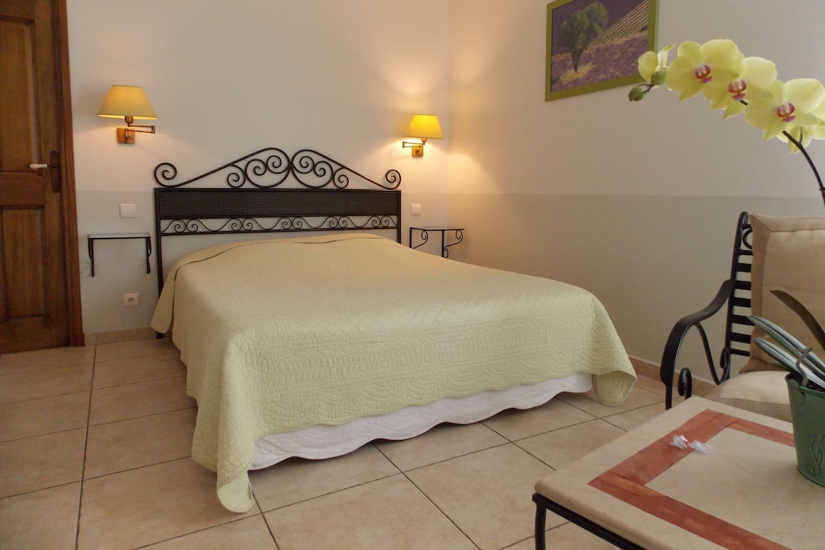 Cama bem arrumada com cabeceira, duas luminárias na parede, um quadro emoldurado e um vaso com flores sobre uma mesa.