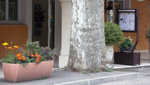 Een hotelingang met een bord waarop 'HOTEL Au Relais Provencal' staat, een grote bloemenplanter en een prikbord met informatie.