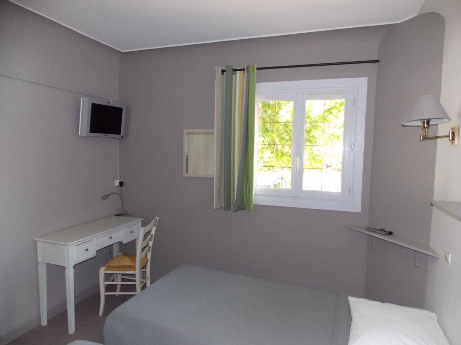 A bedroom with a single bed, a desk with a chair, a television mounted on the wall, a window with curtains, and a shelf with a lamp.